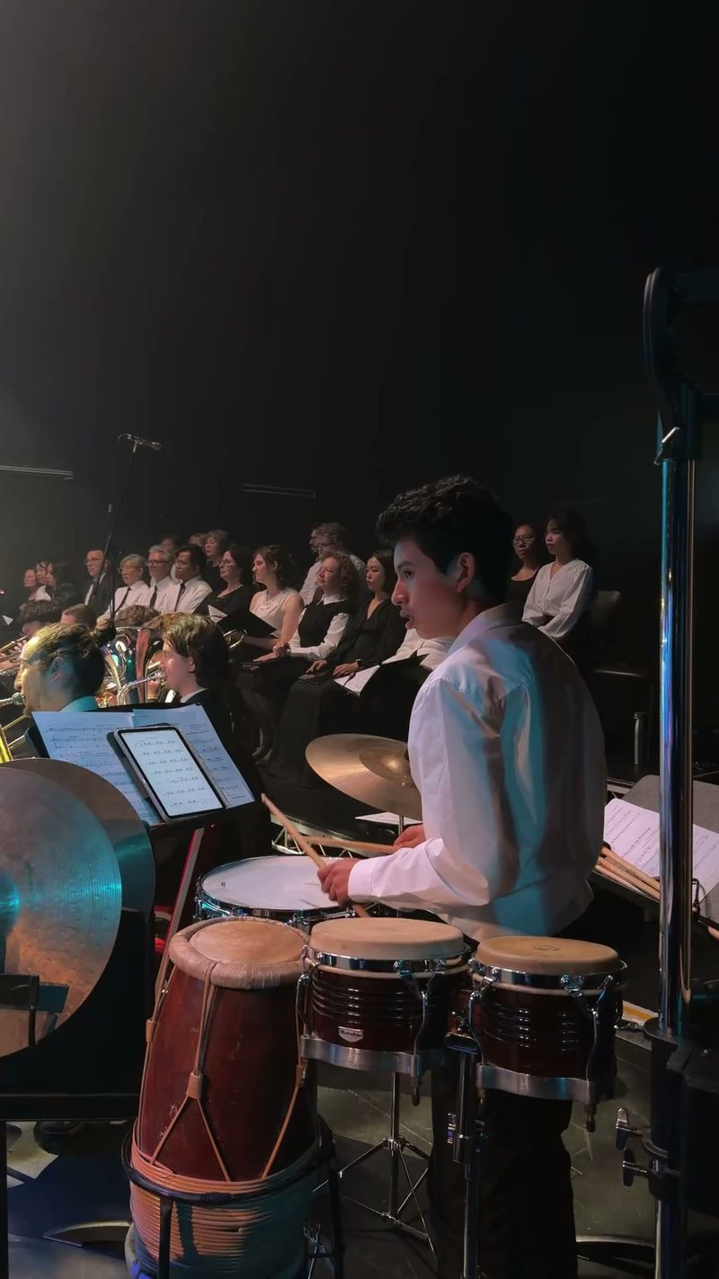 A young percussionist plays drums while the choir performs in the background.