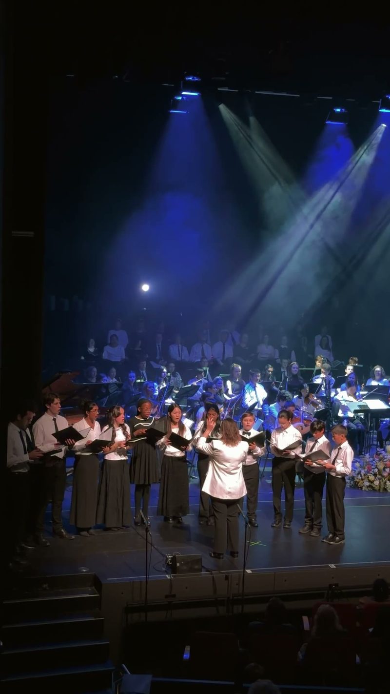 A choir performance with a conductor and orchestral musicians in the background.