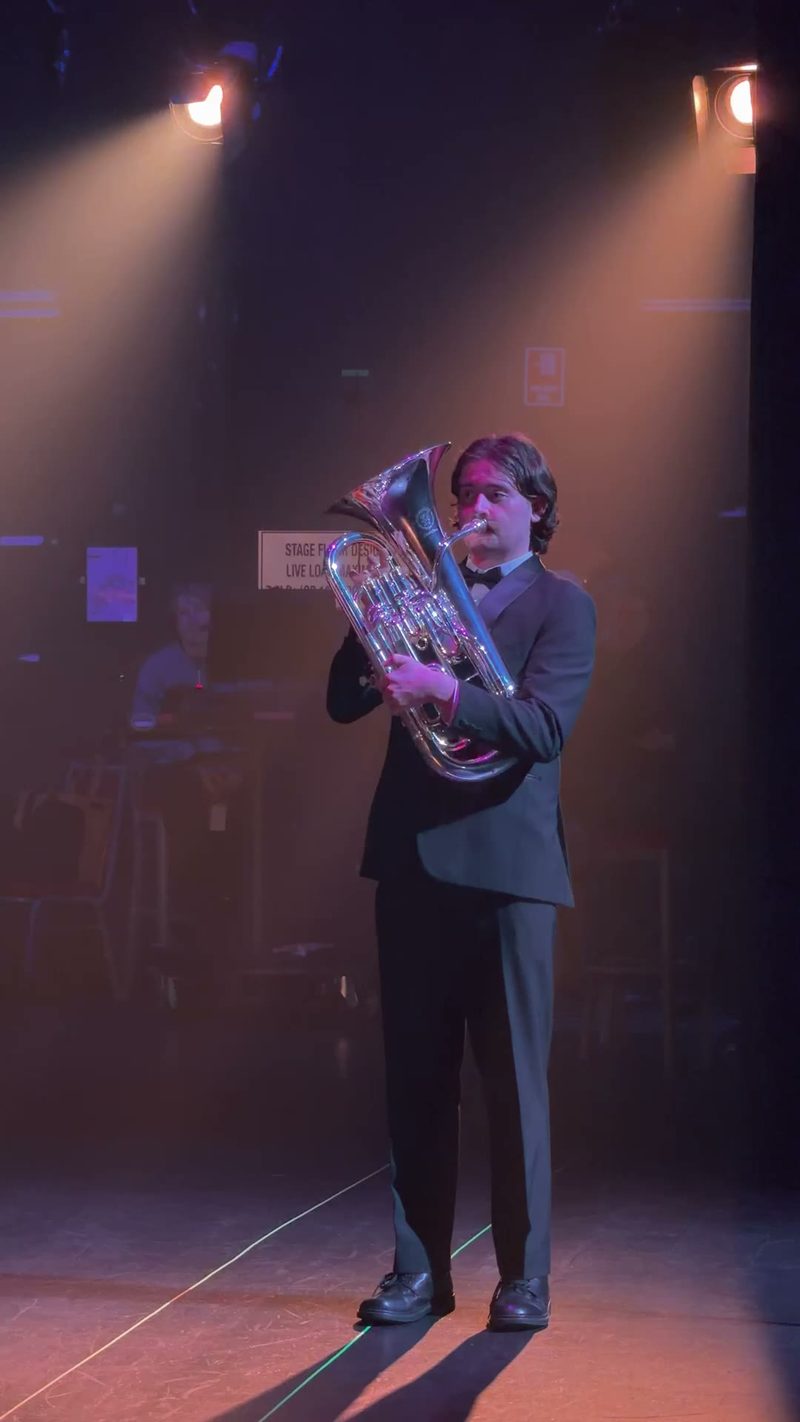 A tuba player performs on stage under dramatic lighting.