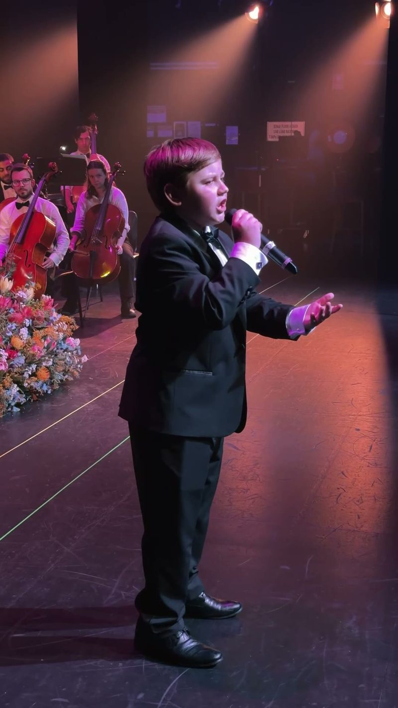 A young boy in a suit sings into a microphone on stage with an orchestra in the background.