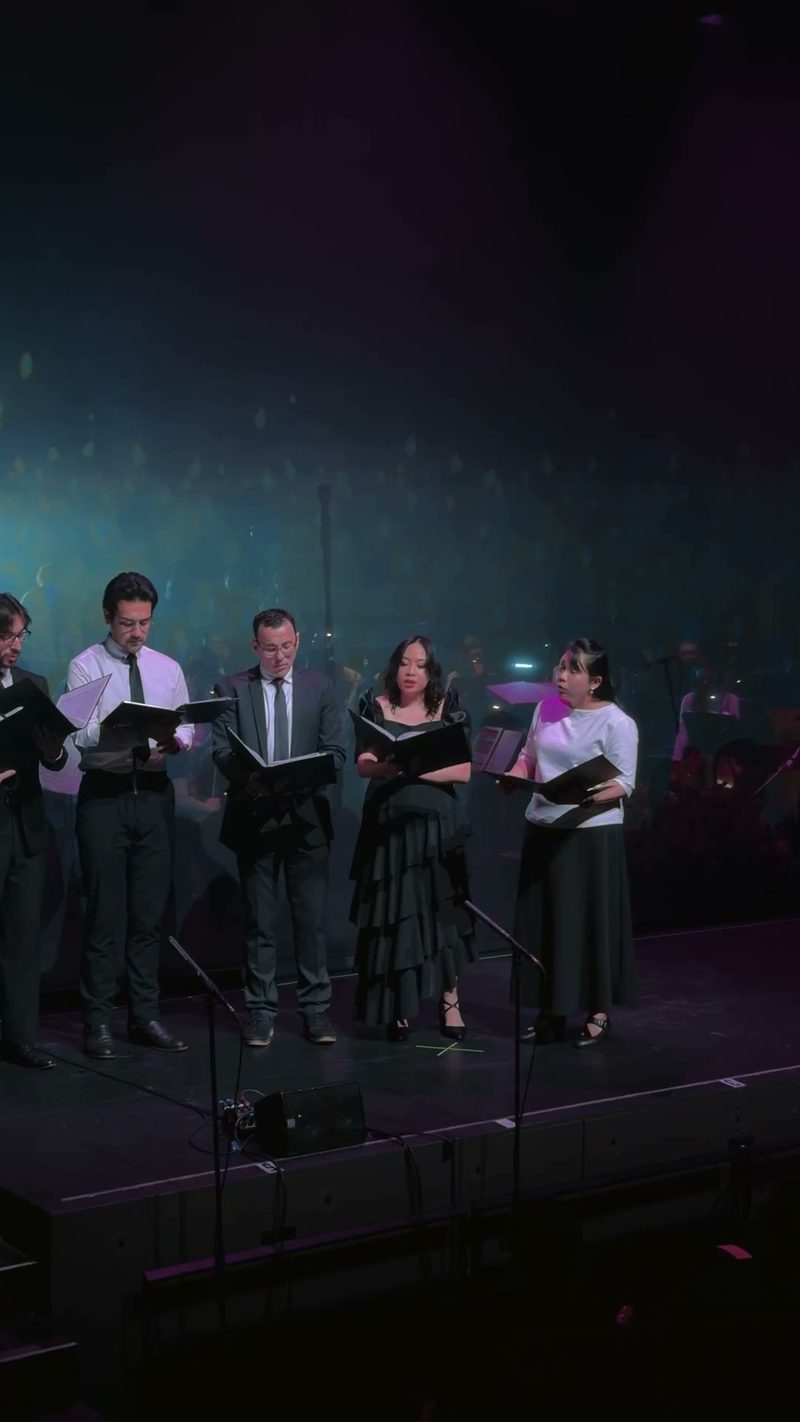 A vocal ensemble performs on stage with sheet music in hand.