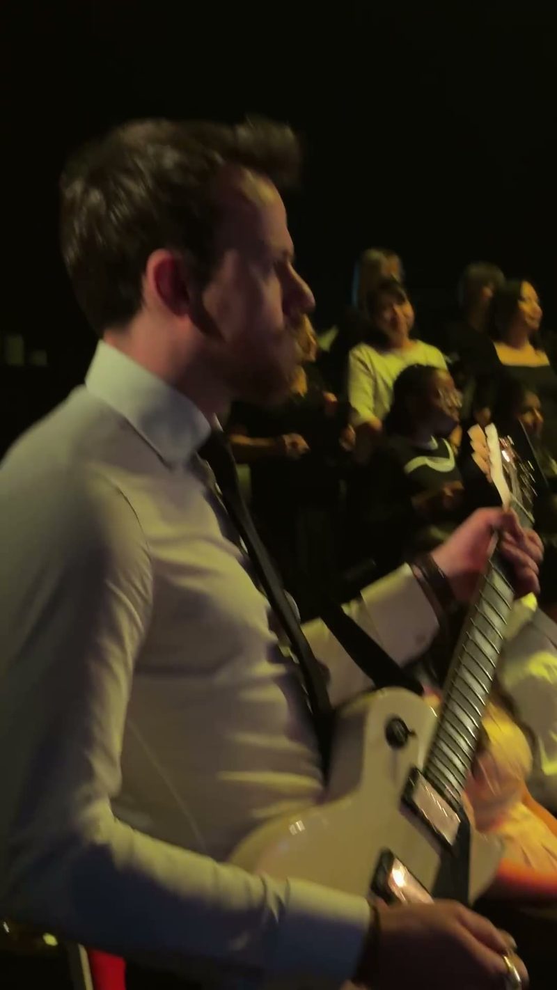 A guitarist dressed in a formal shirt performs on stage while an audience enjoys the music.