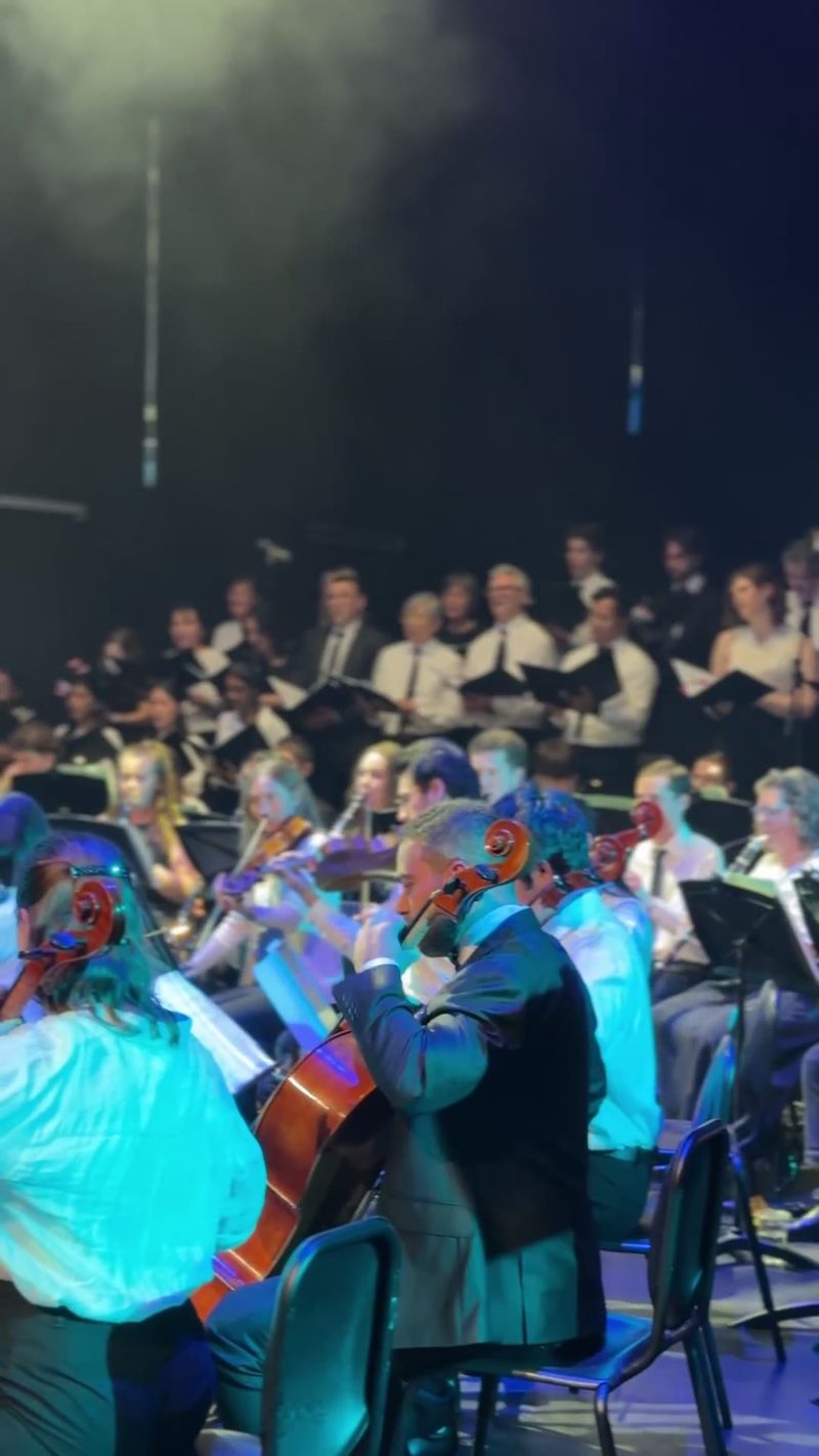 An orchestra performing live with a choir in the background.