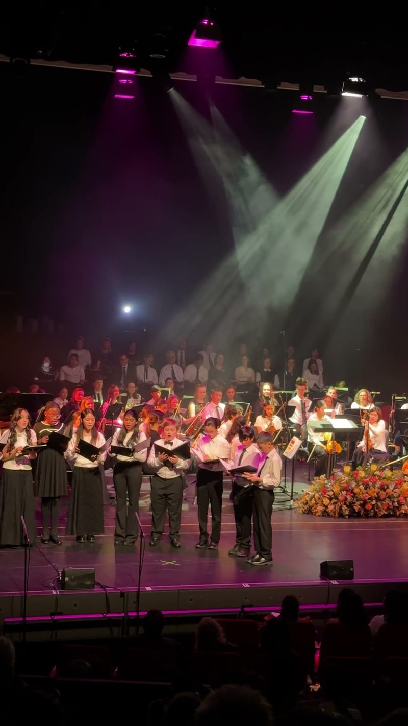 A choir performing on stage with orchestral musicians in the background, illuminated by stage lights.