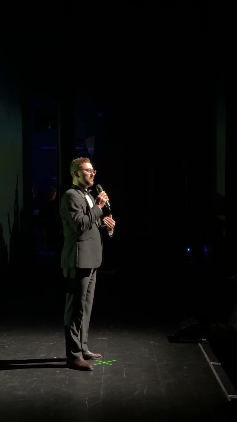 A speaker in formal attire delivering a speech on stage with focused lighting.