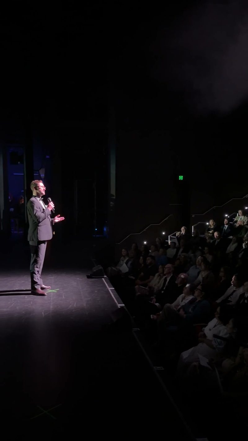 A speaker addresses a crowd from the stage, with audience members holding their phones up in the background.
