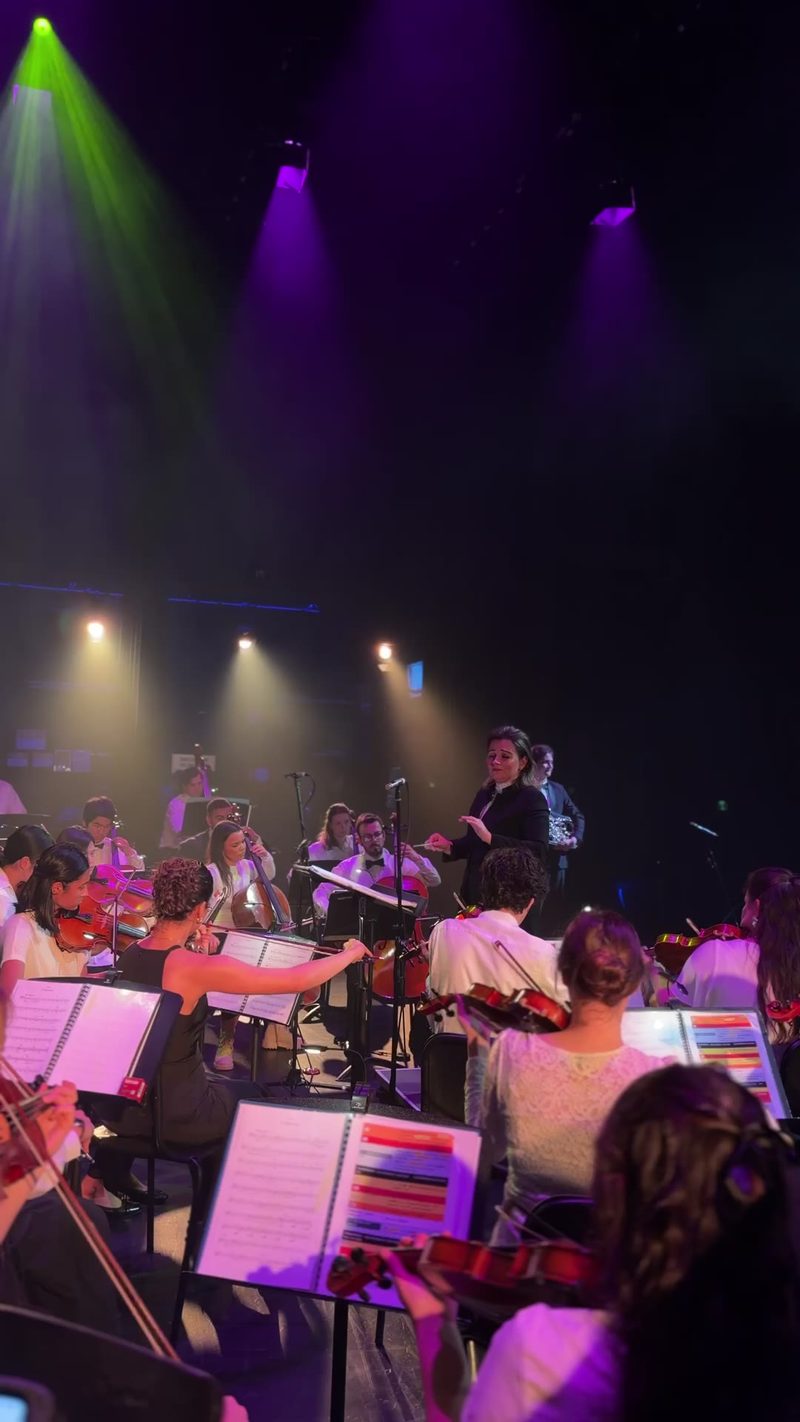 An orchestral performance featuring musicians playing string instruments under colorful stage lighting.