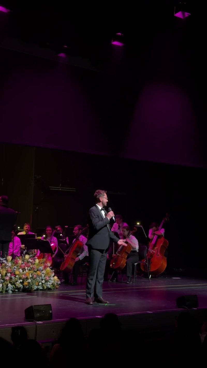 A man speaking into a microphone on stage with an orchestra in the background.