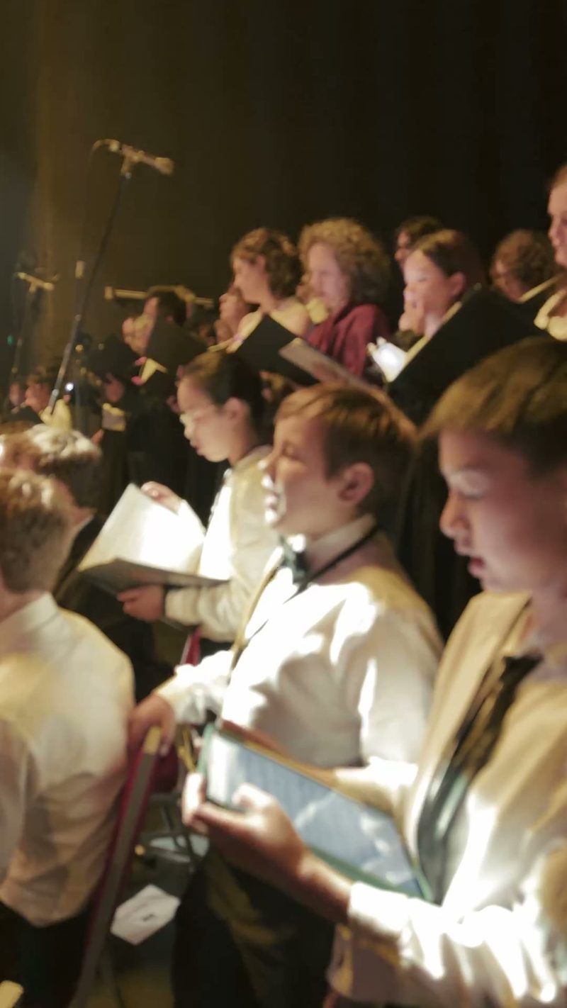 A choir performing on stage with sheet music in hand, illuminated by stage lights.