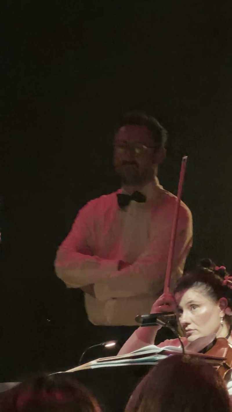 A violinist focuses on her music while a figure in formal attire observes in the background.