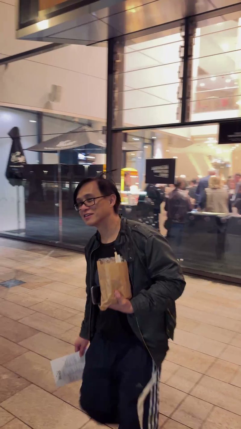 A person holding a bag and a piece of paper appears to be walking outside a venue.