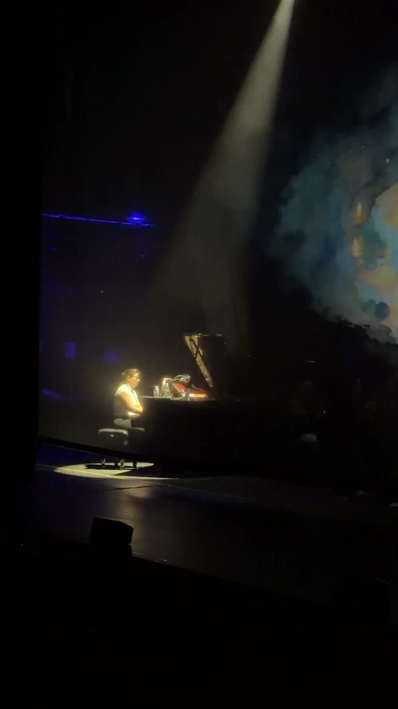 A performer plays a grand piano under a spotlight on stage.
