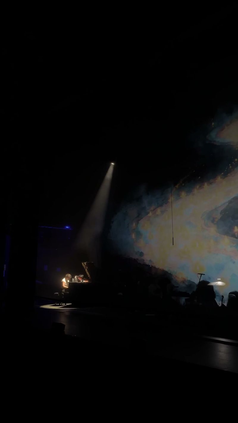 A performer plays the piano under dramatic lighting with a colorful backdrop.