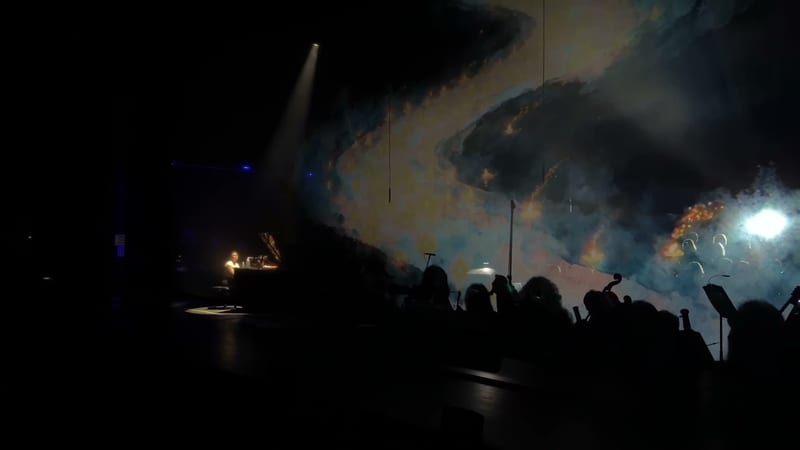 A performer plays a grand piano with an illuminated backdrop during a symphonic performance.