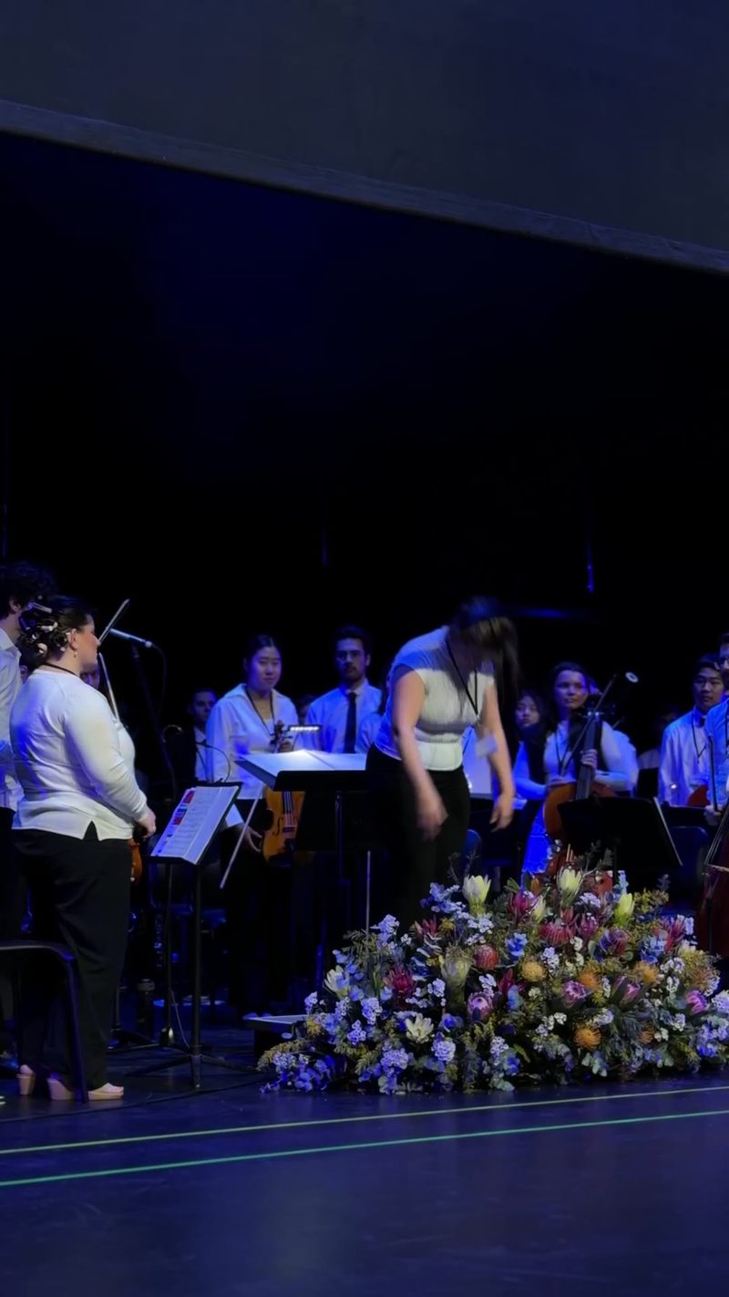 A performance featuring musicians in a symphony orchestra setting, with a floral arrangement in the foreground.