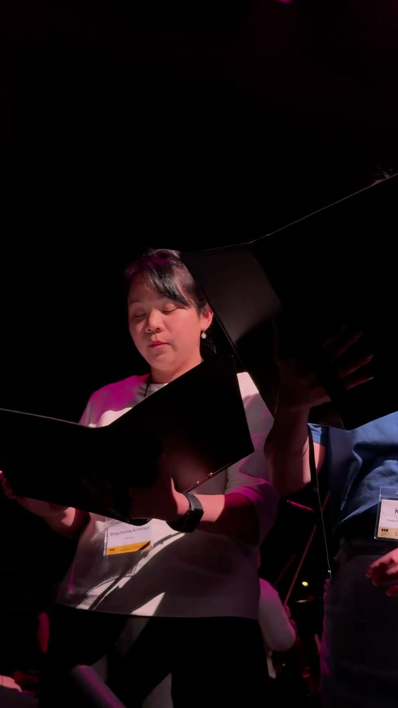 A choir member is focused while reading from a music folder under stage lighting.