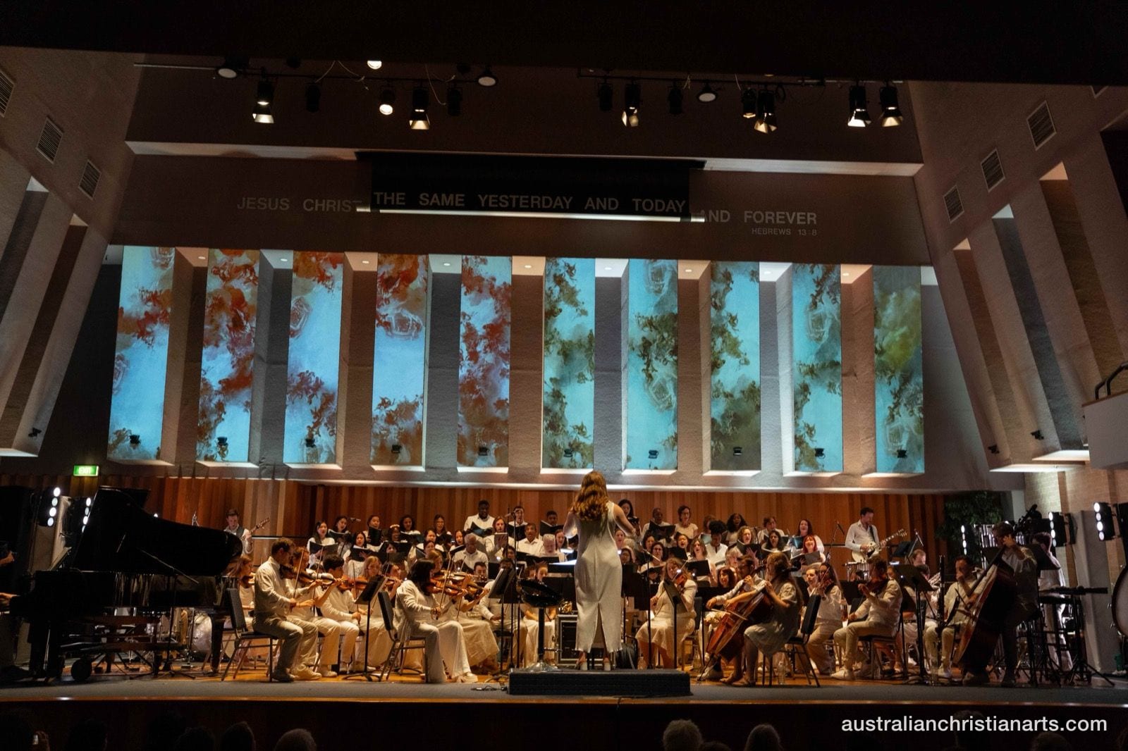 A conductor leads a full orchestra and choir on a church stage illuminated by teal and earth-toned projected panels, with the scripture 'Jesus Christ the same yesterday and today and forever' displayed above.