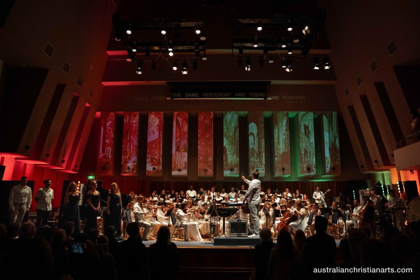 A conductor leads a full orchestra and choir on a dramatically lit stage with projected Renaissance architectural imagery in red and green.