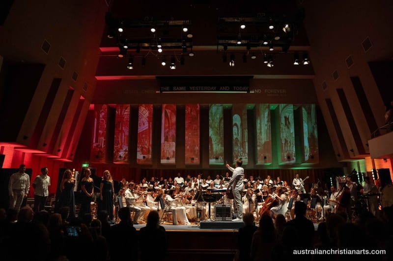 A conductor leads a full orchestra and choir on a dramatically lit stage with projected Renaissance architectural imagery in red and green.
