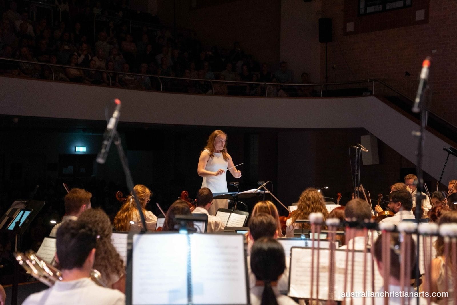 A female conductor in a white dress leads a full symphony orchestra during a concert in a large auditorium packed with audience members in the balcony.