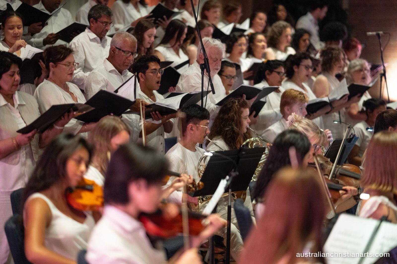 A large choir in white attire sings from black folders while string and brass players perform in the foreground during a concert.