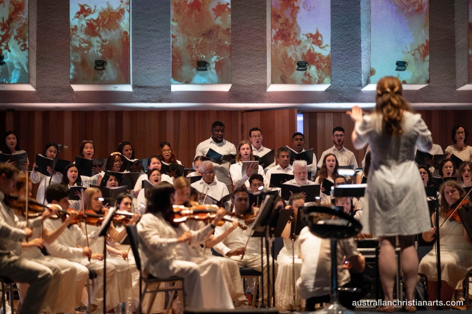 A conductor in a white dress leads a large choir and string orchestra performing in a concert hall with colourful projected artwork on the walls behind them.