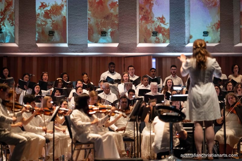 A conductor in a white dress leads a large choir and string orchestra performing in a concert hall with colourful projected artwork on the walls behind them.