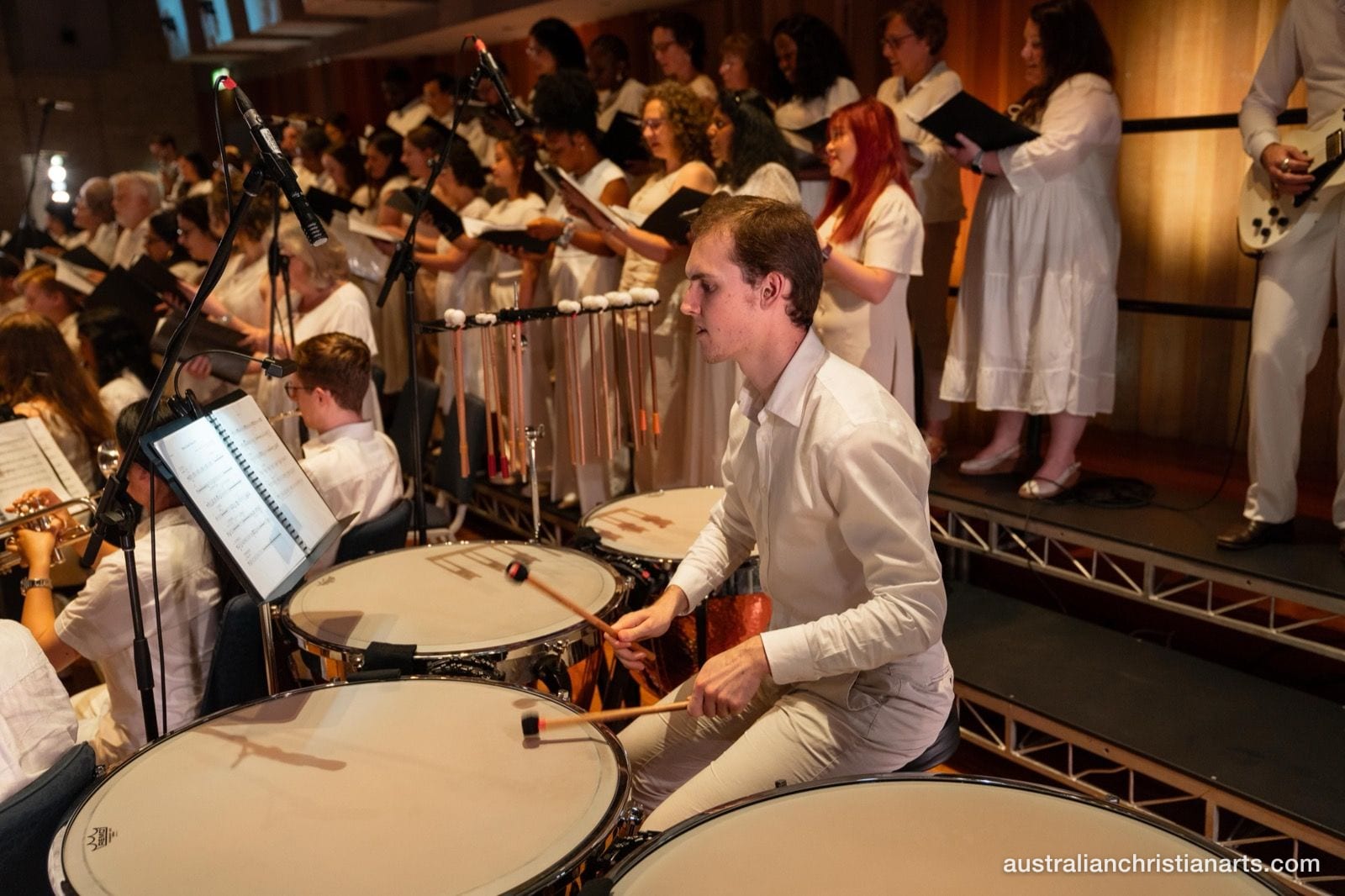 A percussionist in a white shirt plays timpani during a large orchestral and choral concert performance.