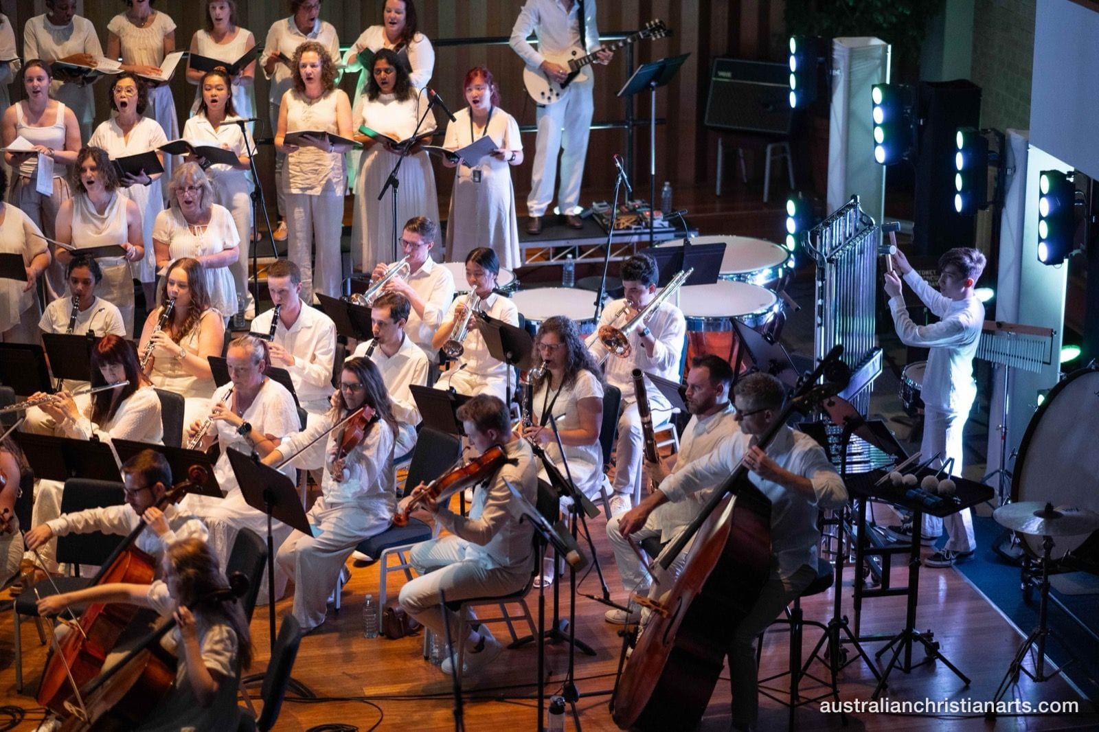 A large ensemble of orchestral musicians and choir singers, all dressed in white, perform together on a concert hall stage with dramatic stage lighting.