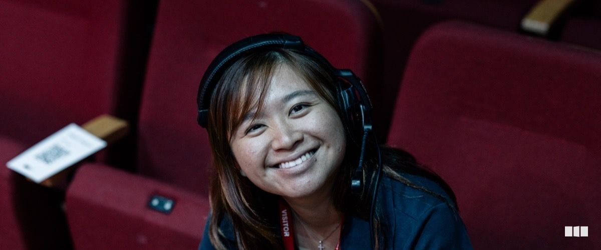A smiling young woman wearing headphones and a visitor lanyard sits among red auditorium seats