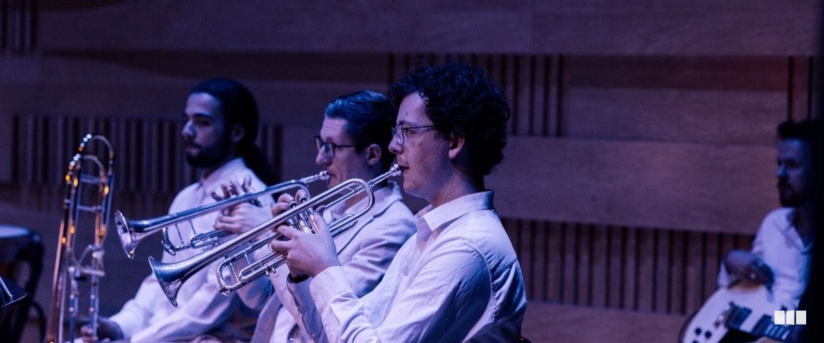 Three trumpet players in white shirts perform together in a warmly lit concert hall with wooden acoustic panelling.