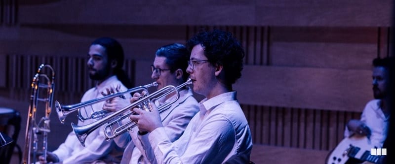 Three trumpet players in white shirts perform together in a warmly lit concert hall with wooden acoustic panelling.