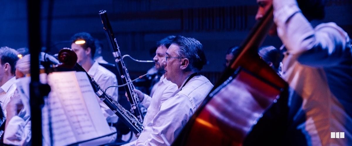 Orchestra musicians perform on stage, with a bassoonist in focus reading from sheet music alongside string and woodwind players under blue-toned stage lighting.