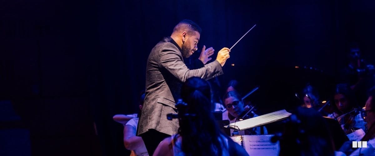 An orchestra conductor leads the ensemble with baton raised under dramatic blue stage lighting