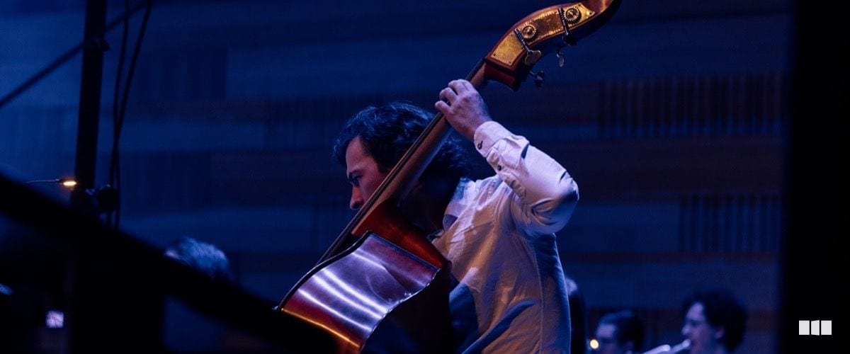 A double bass player performs with intensity on a dimly lit stage bathed in blue and warm light.