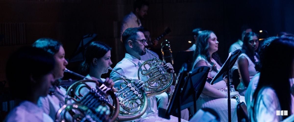French horn players in white attire perform together during an orchestra concert under blue-tinted stage lighting.