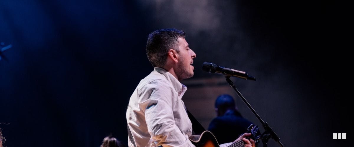 A male vocalist in a white shirt performs at a microphone while playing acoustic guitar on a darkly lit stage