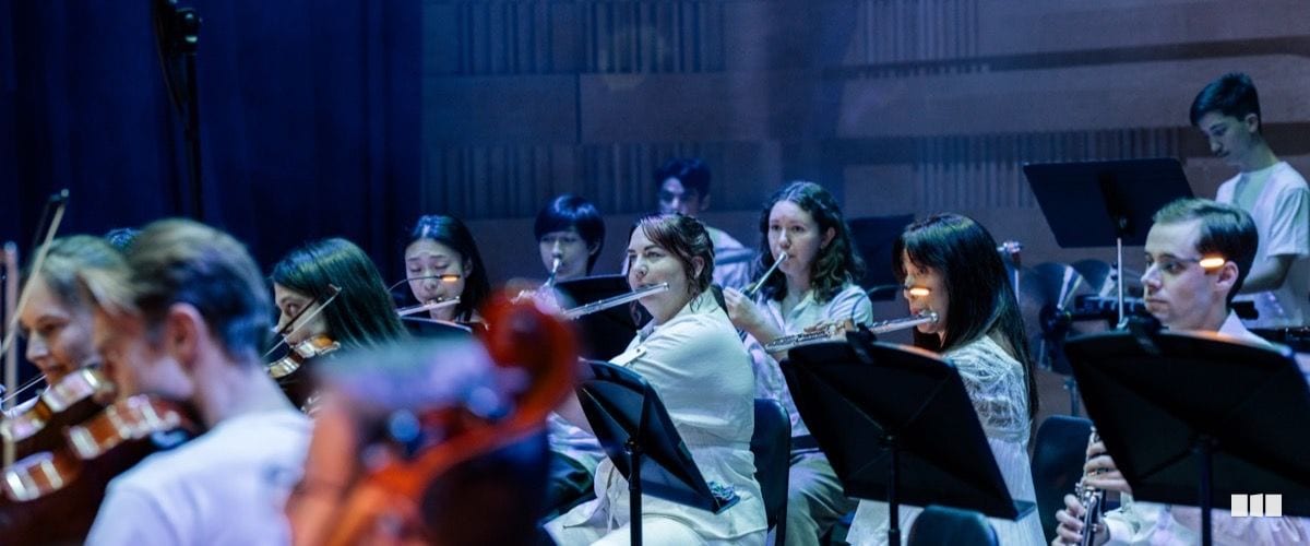 Flute and string players perform together on stage during an orchestral concert, reading from music stands under blue stage lighting.