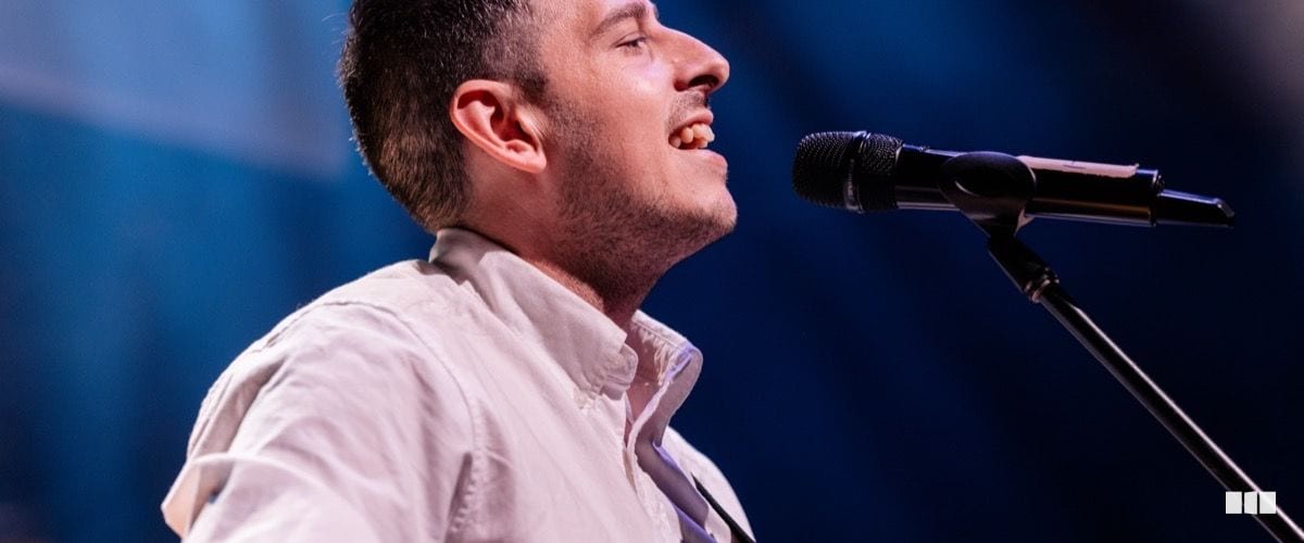 A male vocalist performs with passion at a microphone stand on a concert stage, bathed in blue light.