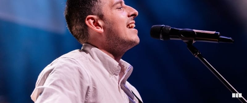 A male vocalist performs with passion at a microphone stand on a concert stage, bathed in blue light.
