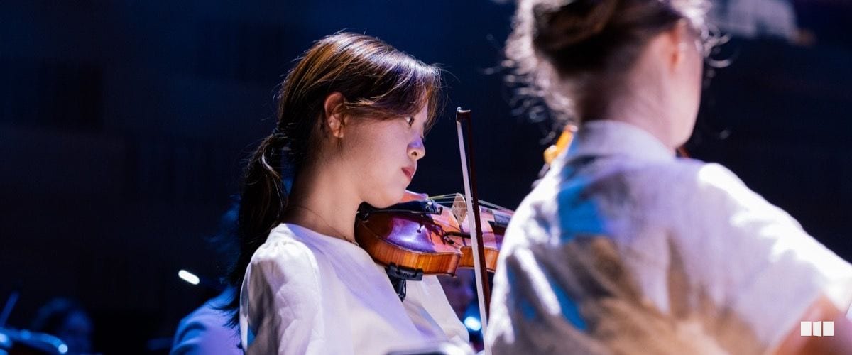A young woman in a white top plays violin during an orchestral performance under stage lighting.