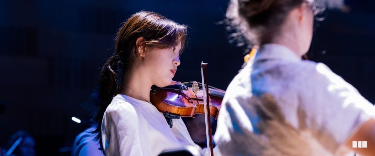 A young female violinist in a white top performs with focused concentration during an orchestral concert under blue stage lighting.