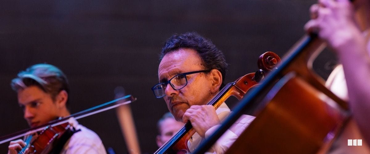 A cellist wearing glasses focuses intently while performing alongside other string players in an orchestra concert.