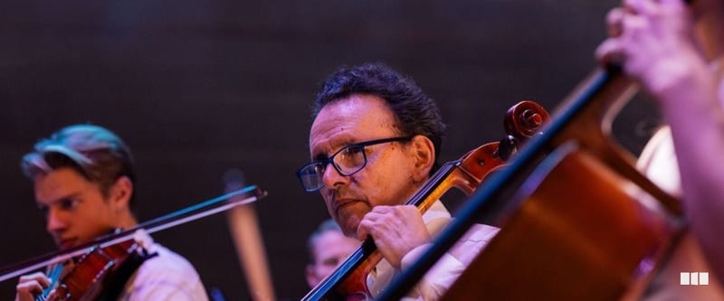A cellist wearing glasses focuses intently while performing alongside other string players in an orchestra concert.