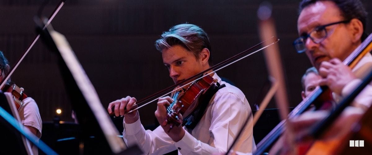 A young violinist performs with focused intensity alongside fellow string players during an orchestral concert.