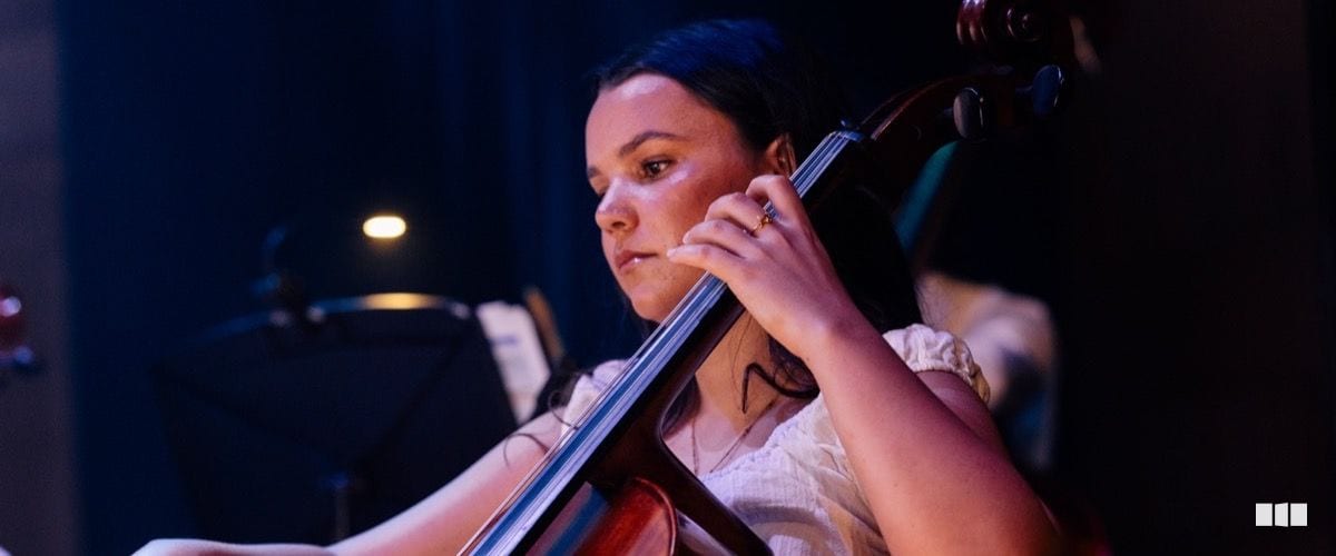 A young woman concentrates intently while playing cello during an orchestra performance under stage lighting