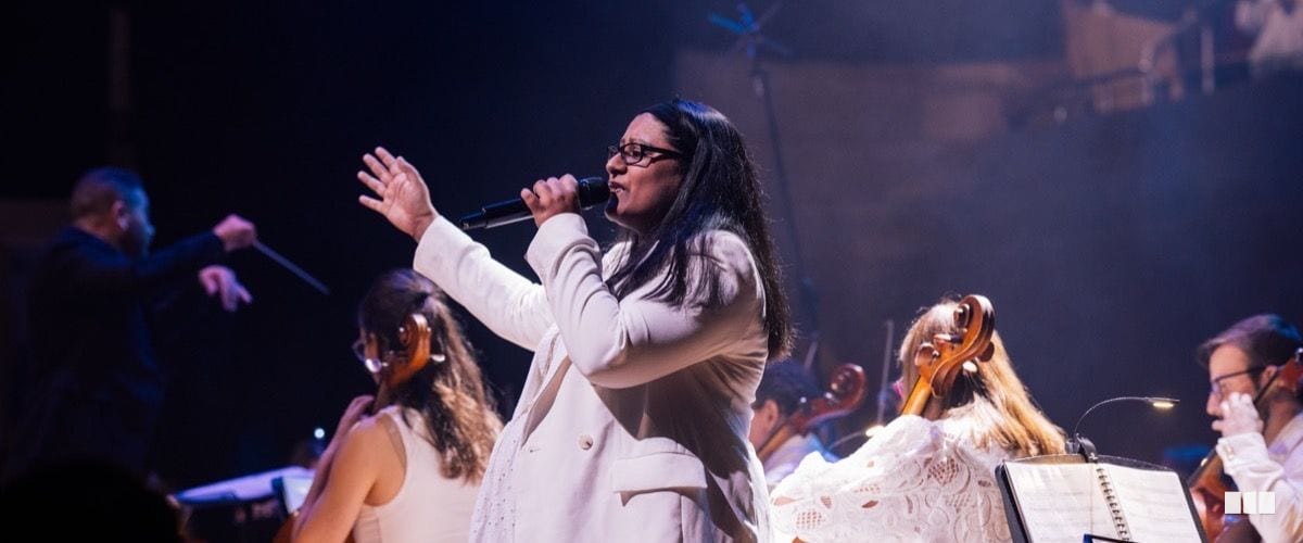 A female soloist in a white suit sings passionately into a microphone while an orchestra performs behind her and a conductor is visible in the background