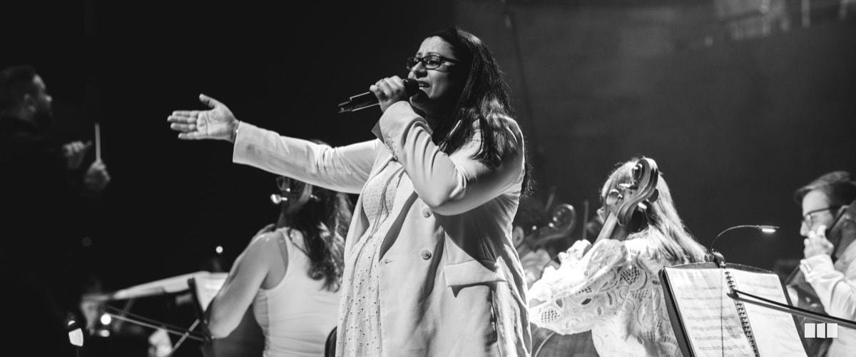A female soloist sings passionately into a microphone with one arm outstretched, backed by orchestra musicians on a darkened stage.