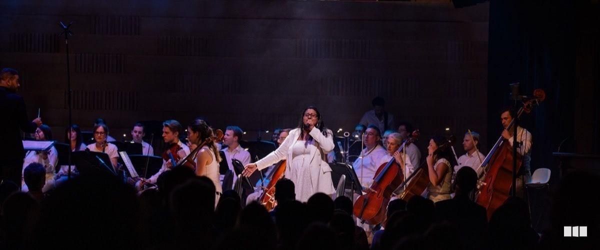 A soloist in white performs with microphone in hand, backed by a full orchestra on a warmly lit concert stage.