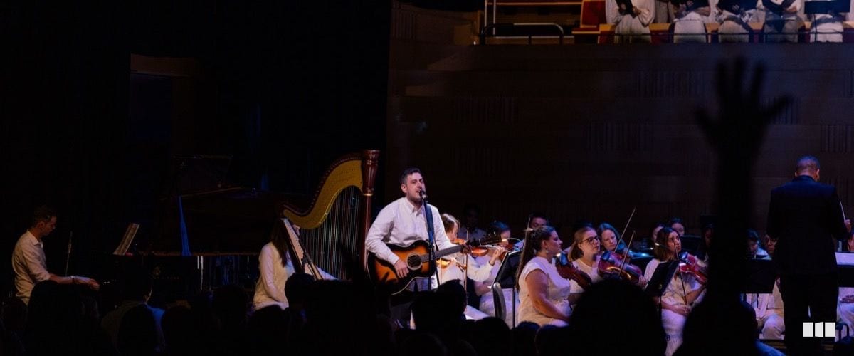 A male soloist plays acoustic guitar and sings into a microphone, accompanied by a full orchestra including harp and strings on a dimly lit concert stage.